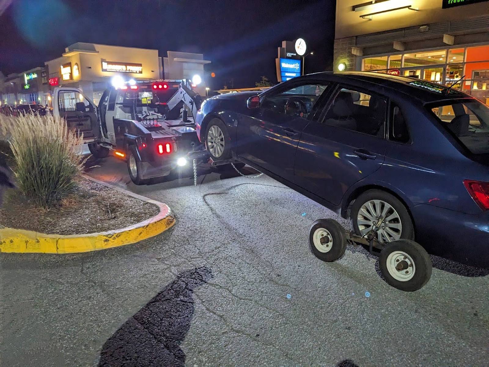Nighttime scene of a tow truck lifting the front wheels of a dark blue sedan in a commercial parking lot, with retail storefronts including Walmart visible in the background.
