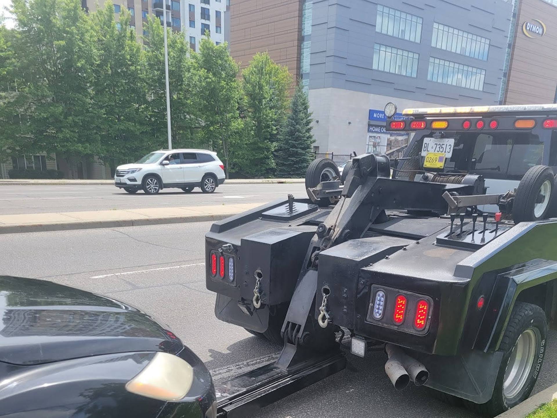 Rear view of a black heavy-duty tow truck towing a black car on a city street, with buildings, trees and a white SUV in the background.