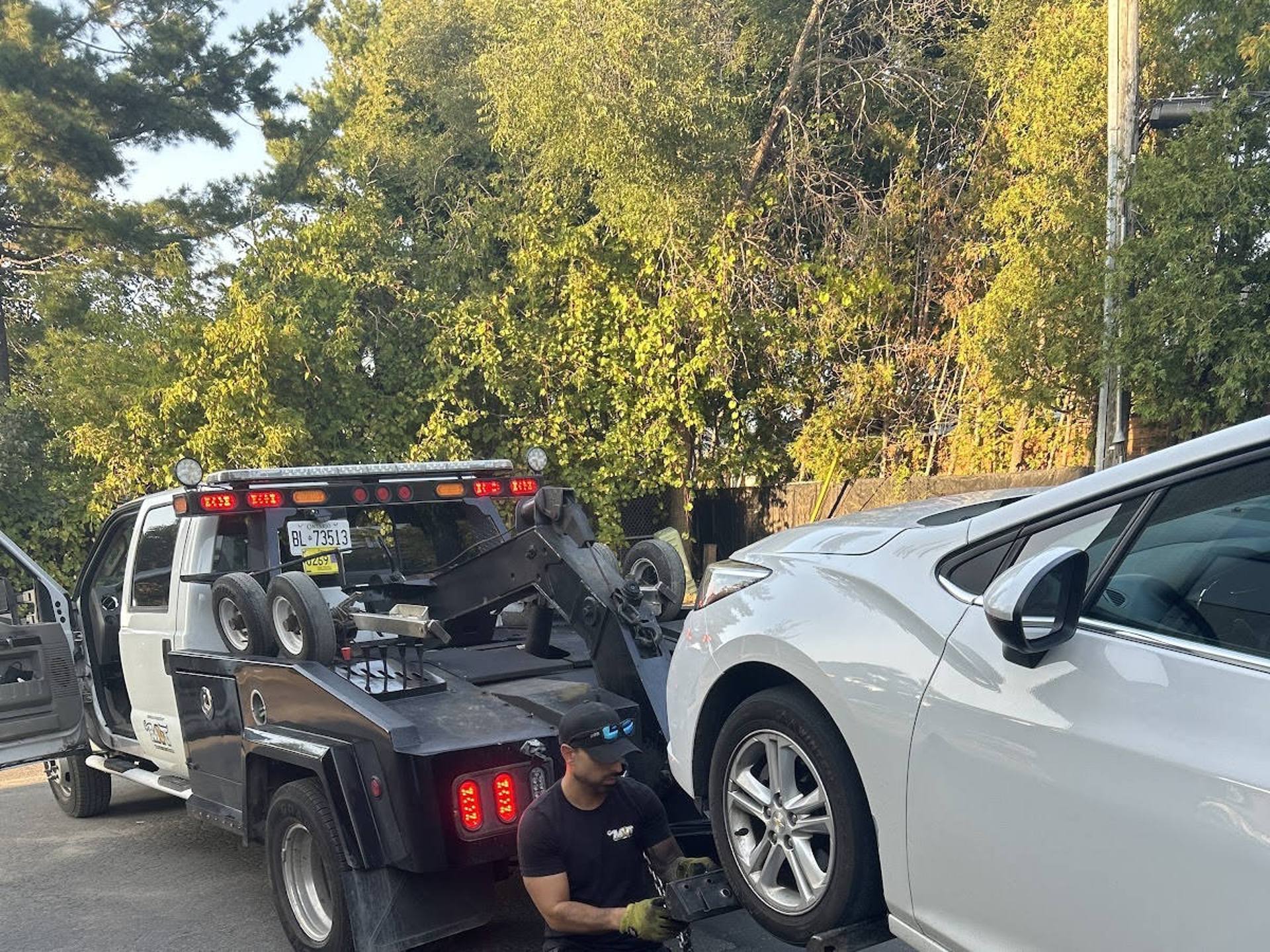 A tow truck operator secures the front wheel of a white sedan to a wheel dolly to load the vehicle onto a flatbed tow truck, with trees in the background on a sunny day.