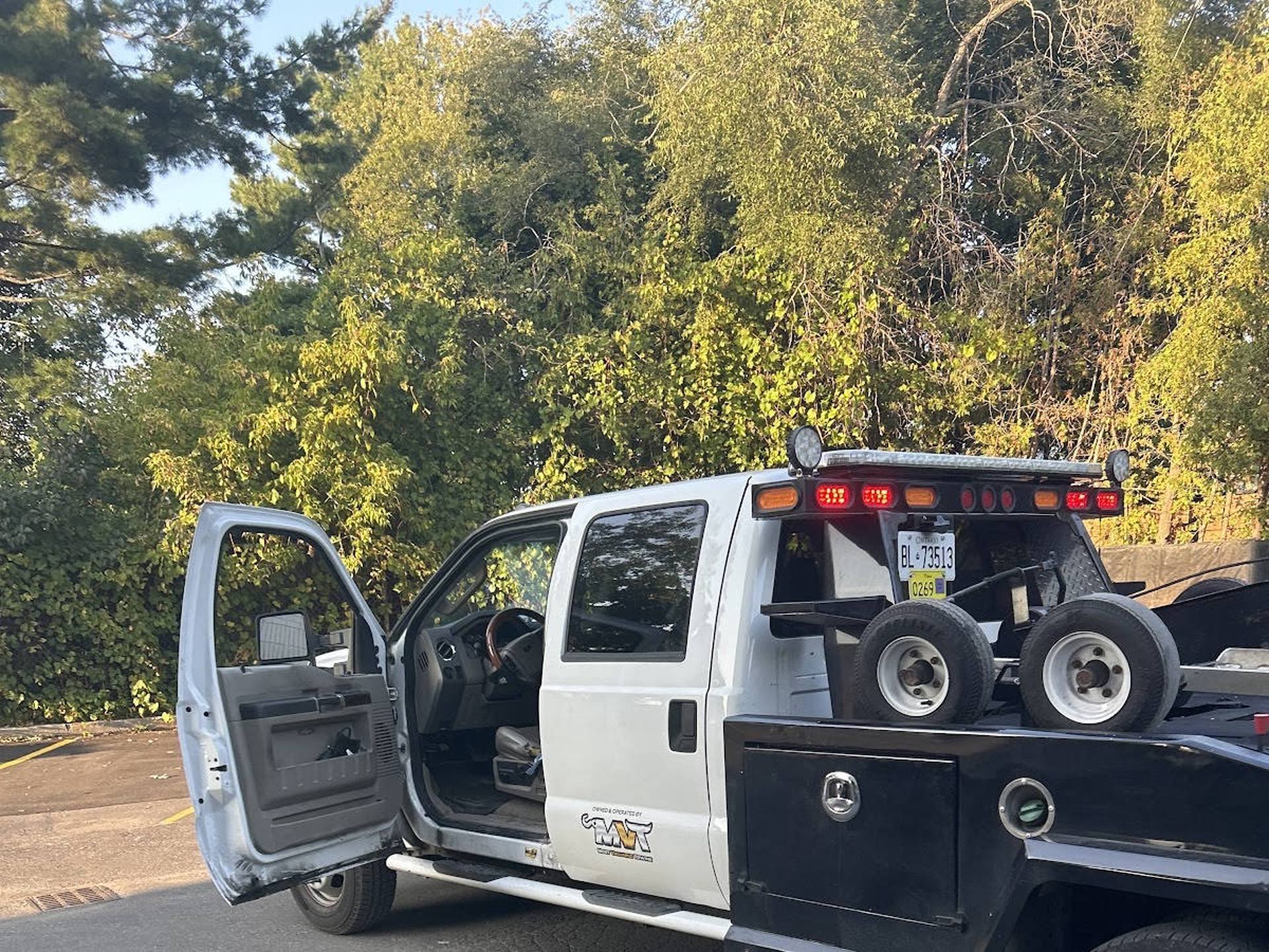 White flatbed tow truck with driver door open, parked in a paved lot with dense green trees in the background, showing rear emergency lights and flatbed with spare tires