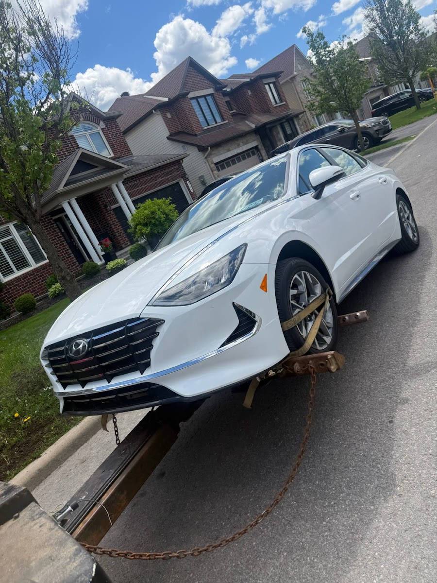 White Hyundai sedan secured on a flatbed tow truck on a residential suburban street, with houses and greenery in the background under a partly cloudy sky