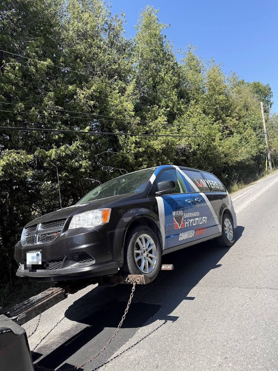 Black branded dealership shuttle minivan secured by chain on flatbed tow truck on rural road