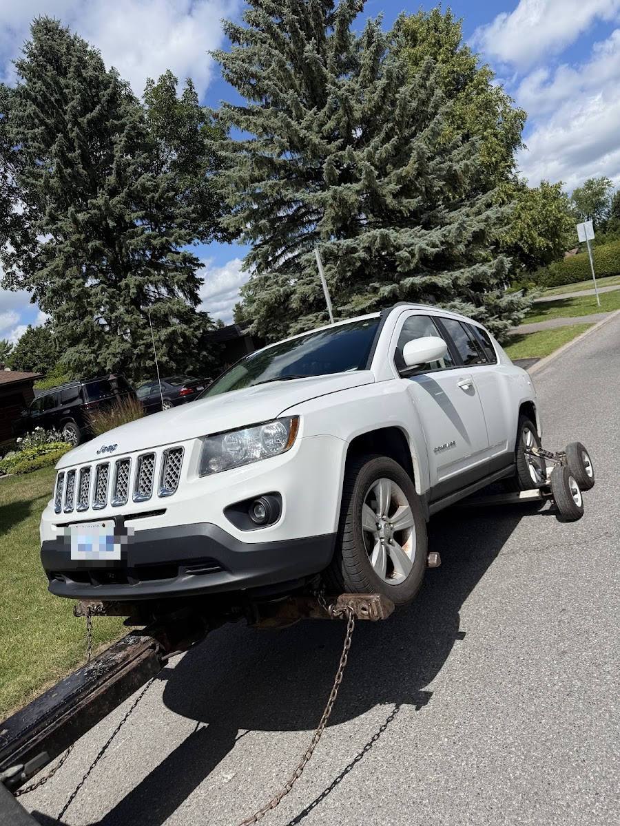 White Jeep Compass on wheel-lift tow truck chains in sunny suburban neighborhood with pine trees