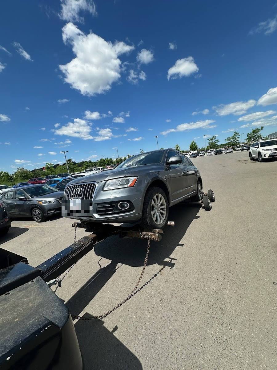 Grey Audi Q5 SUV chained on flatbed tow truck in sunny commercial parking lot