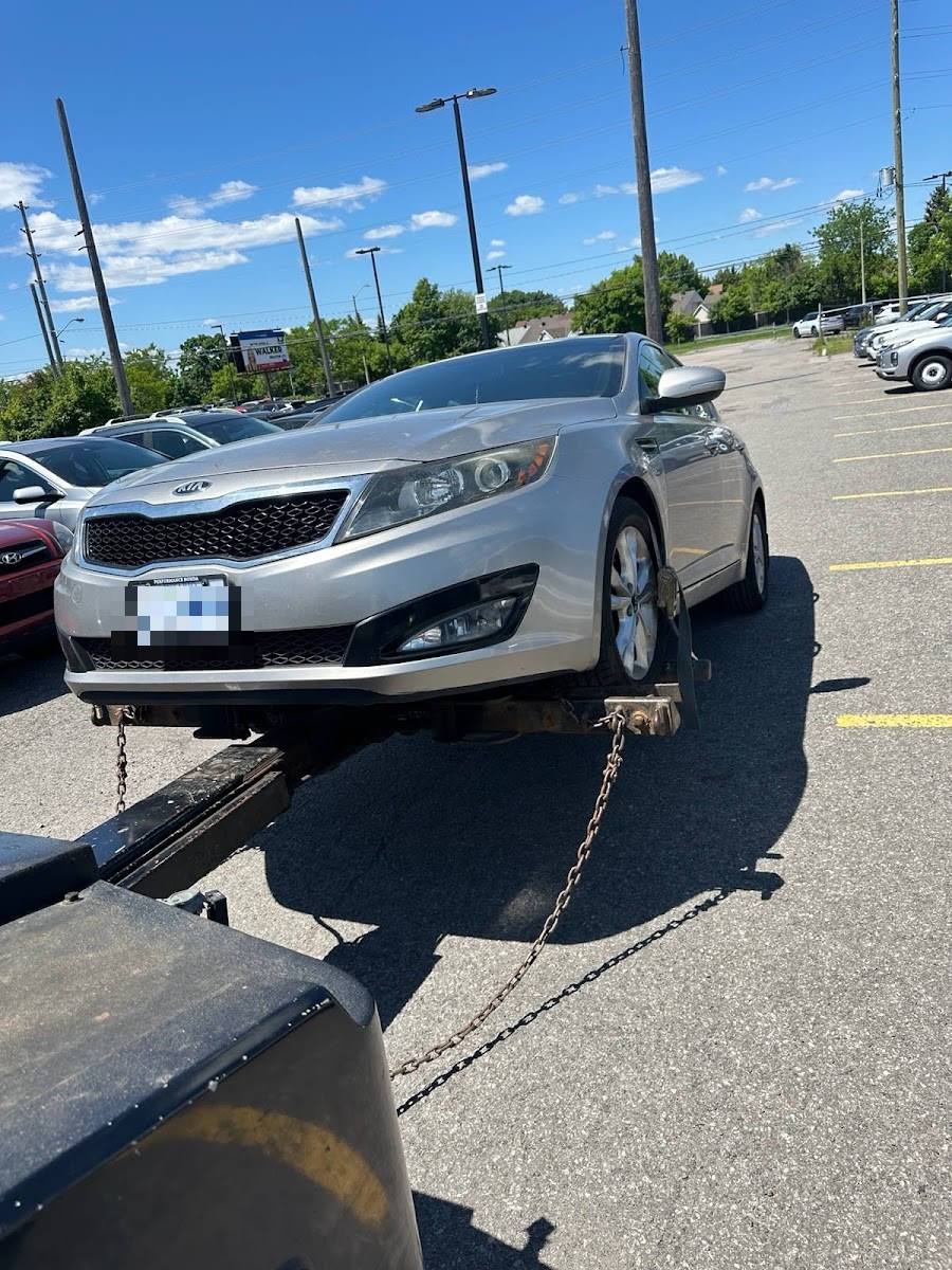 Silver Kia Optima sedan secured with chains on flatbed in sunny commercial parking lot