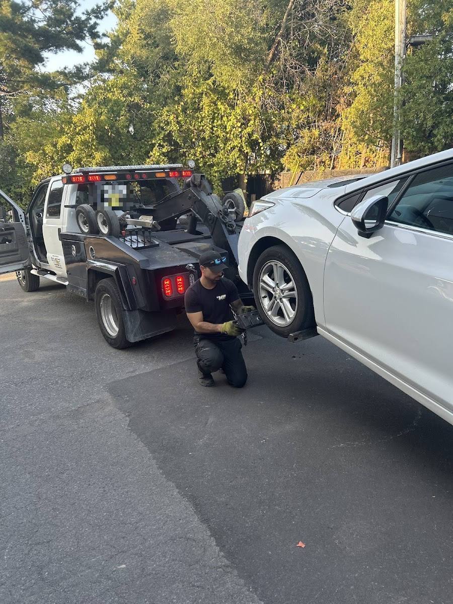 Tow truck operator in black uniform kneeling to hook white sedan beside black wheel-lift truck