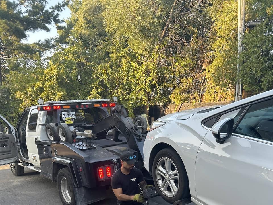 A tow truck operator secures the front wheel of a white sedan to a wheel dolly to load the vehicle onto a flatbed tow truck, with trees in the background on a sunny day.