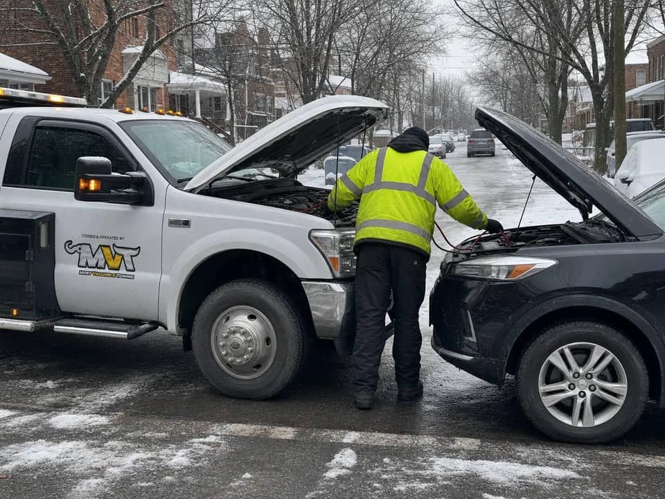 A Most Valuable Towing technician in a hi-vis jacket boosting a dead battery under the open hood of a black SUV, the branded MVT tow truck alongside on a snowy Ottawa street.