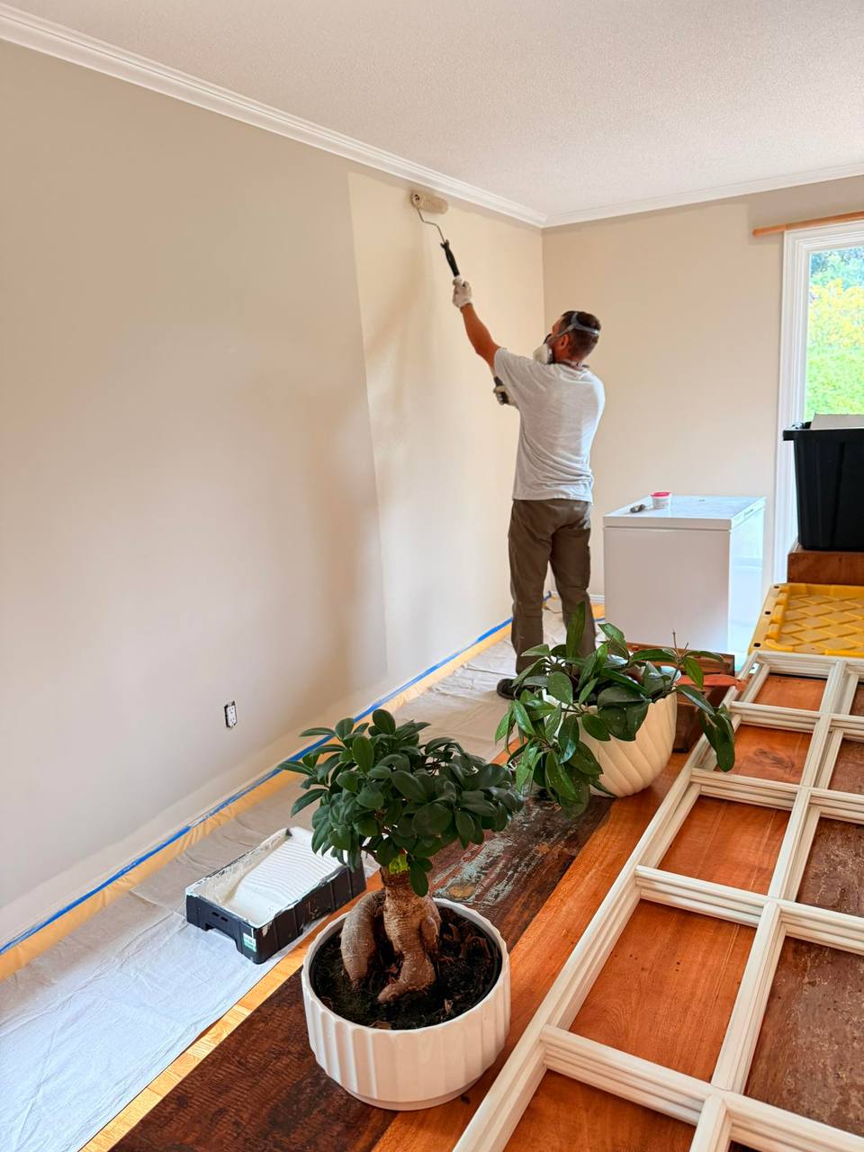 Painter rolling a fresh coat onto an interior wall in a prepped living room, trim removed and floors protected.